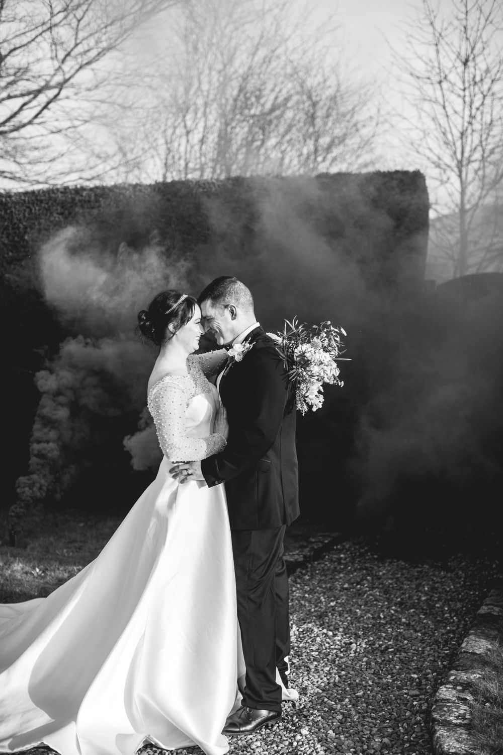 Black and white wedding portrait with dramatic smoke effects and a flowing white dress in a garden setting.