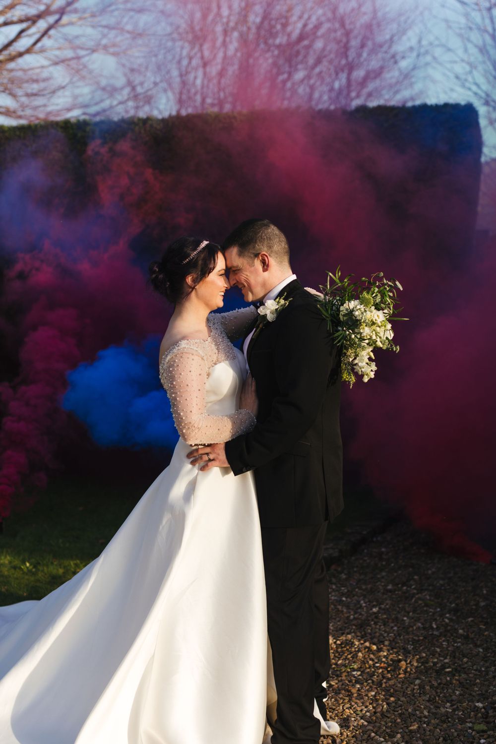 Romantic wedding portrait with vibrant blue and pink smoke effects against an outdoor backdrop.