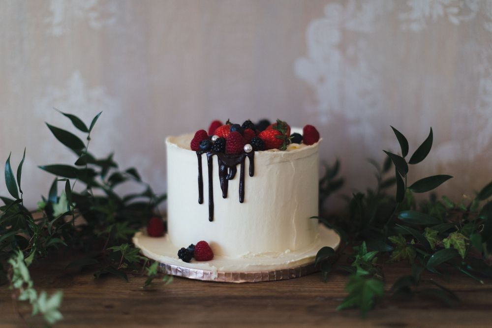 White wedding cake decorated with fresh berries and chocolate drip on wooden table with greenery.