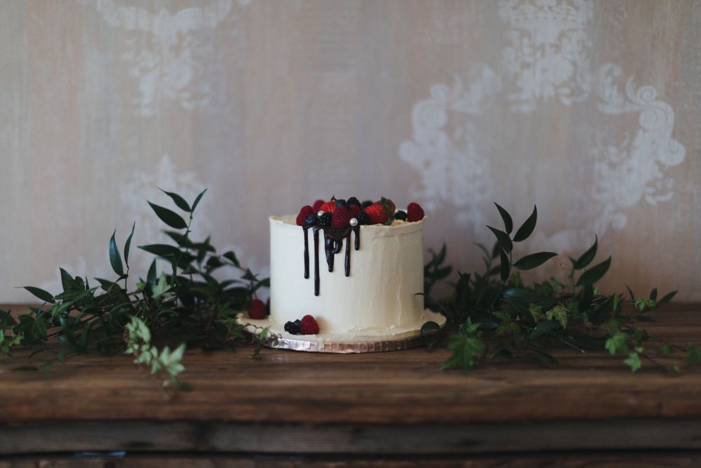 Elegant white cake with dripping chocolate and berries displayed on rustic wooden surface with foliage.