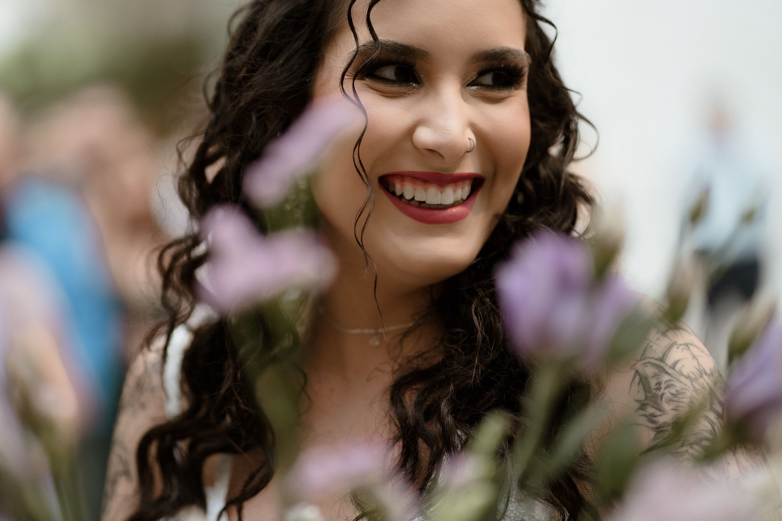 A bride with dark curly hair and natural makeup smiles through purple flowers in soft focus.