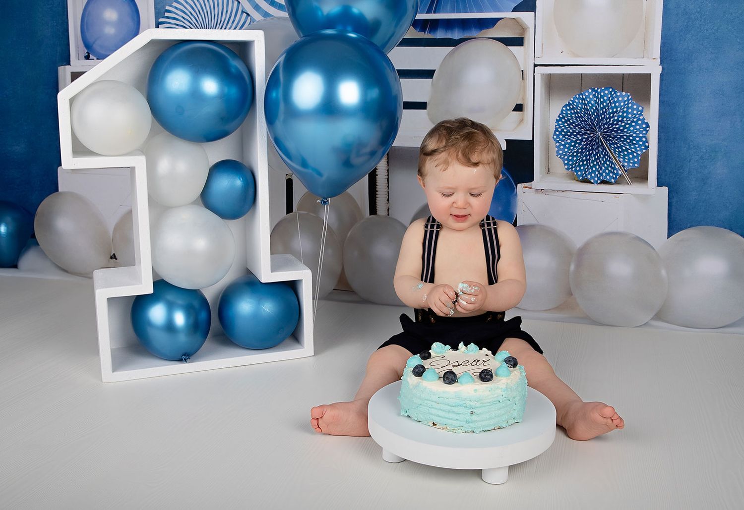First birthday setup with baby seated by cake surrounded by blue and white balloons.