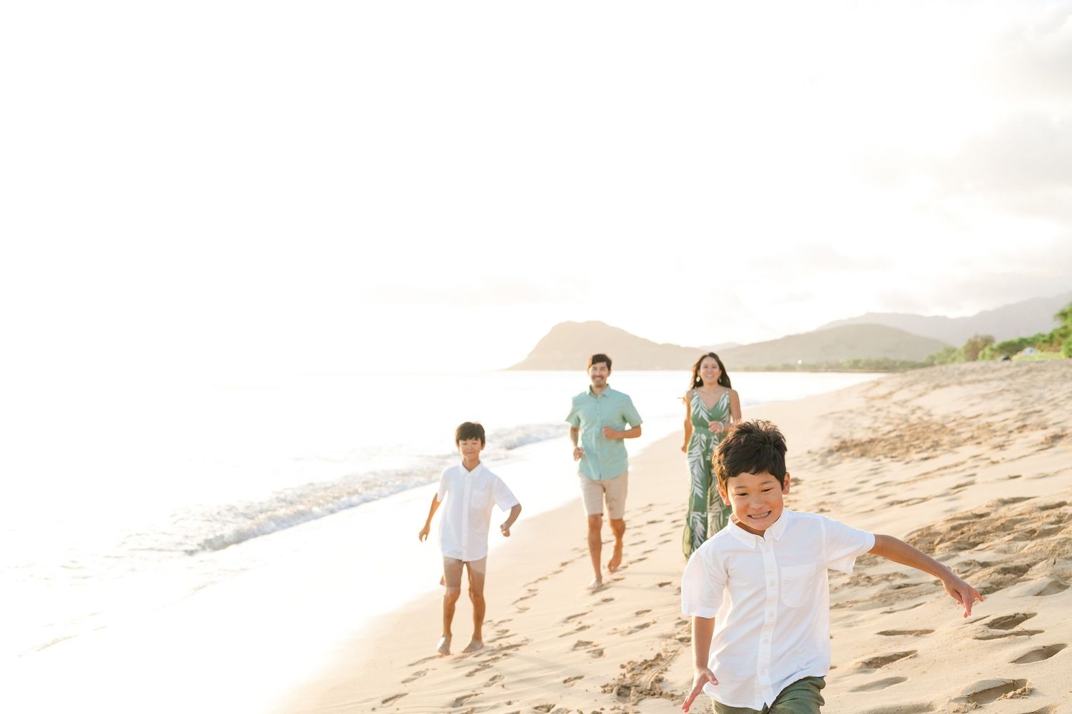 A family walks together along a scenic beach at sunset with mountains in the background. Oahu Family Photographer