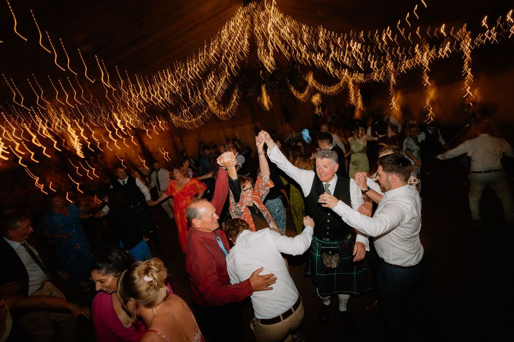 Festive wedding reception with guests dancing under warm string light decorations.