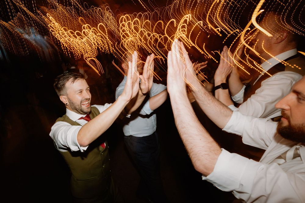 Wedding guests dance together under warm string lights creating swirling light trails in this long exposure shot.