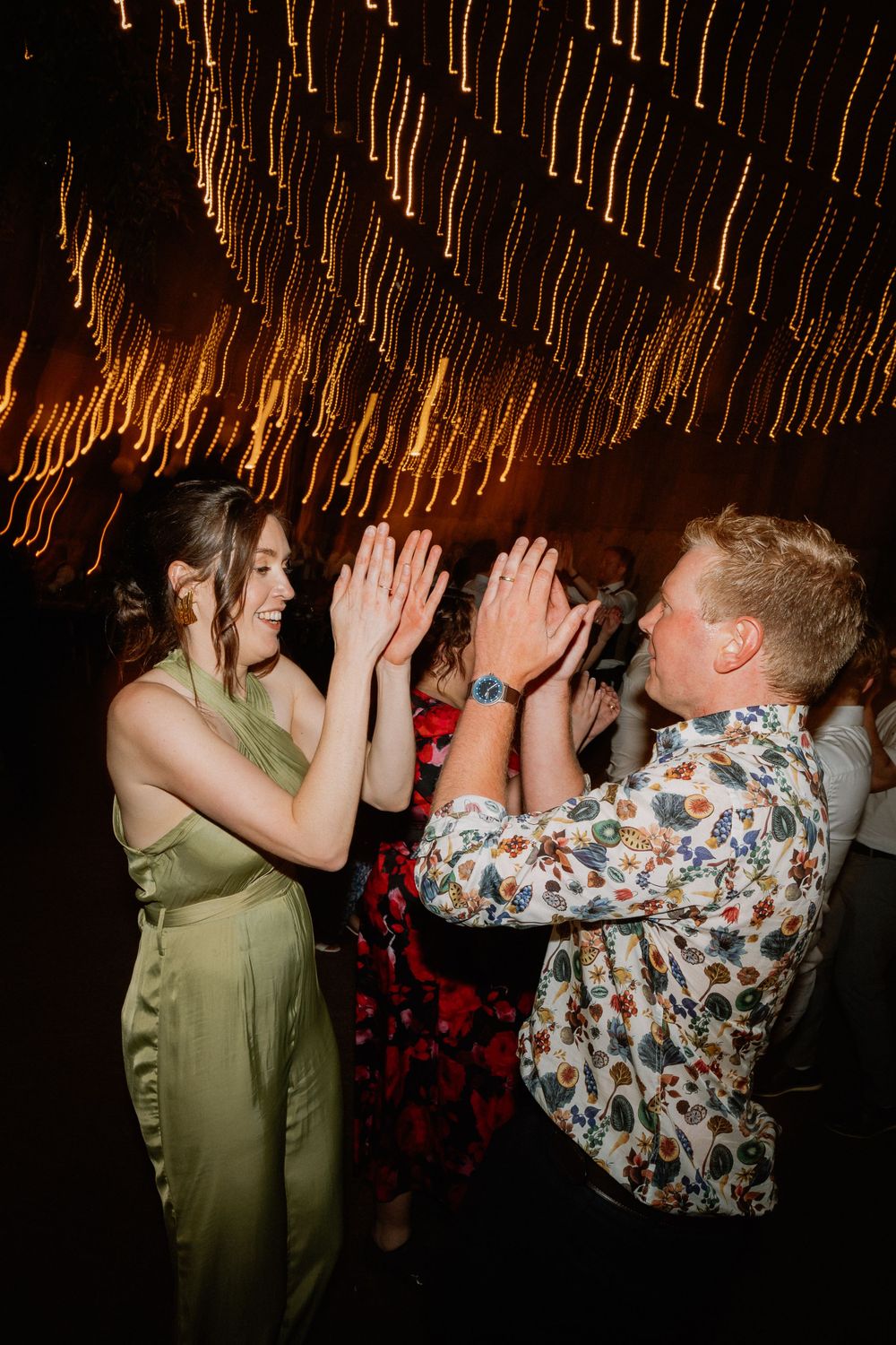 Party guests clapping hands while dancing under glowing string light decorations.