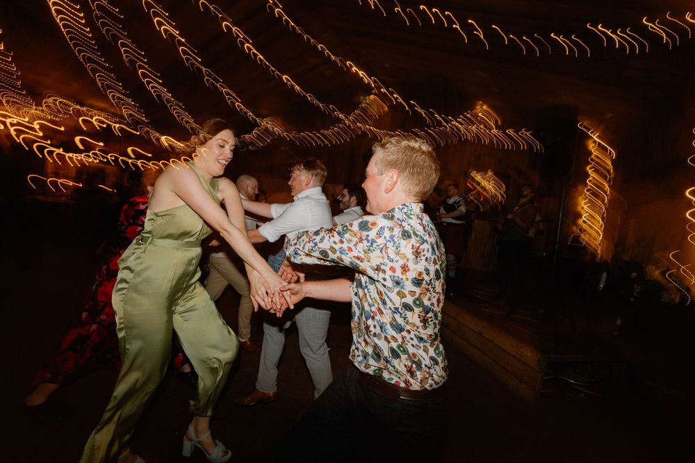 Wedding guests dancing together under decorative string lights at an indoor reception.