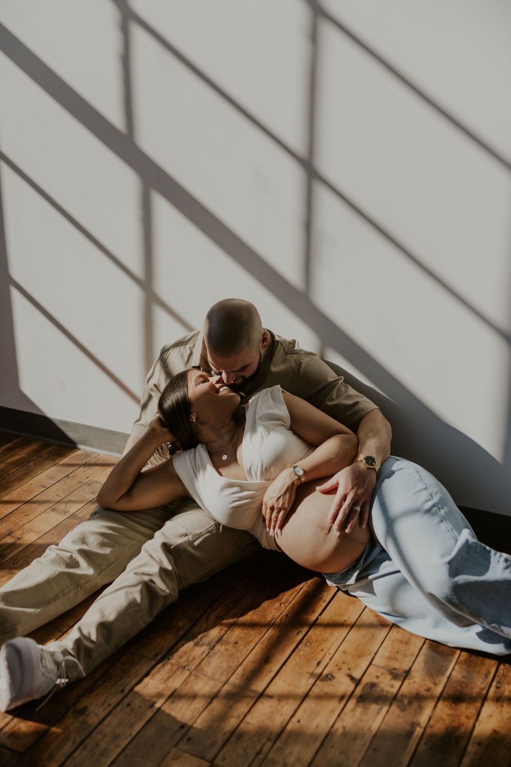 A couple in casual clothing lies on a wooden floor with dramatic shadows from window blinds creating stripes across them.