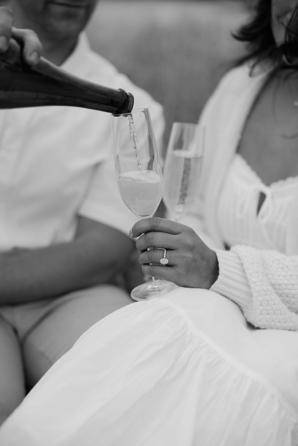 Black and white photo of champagne being poured into a glass held by someone wearing white attire with a ring.