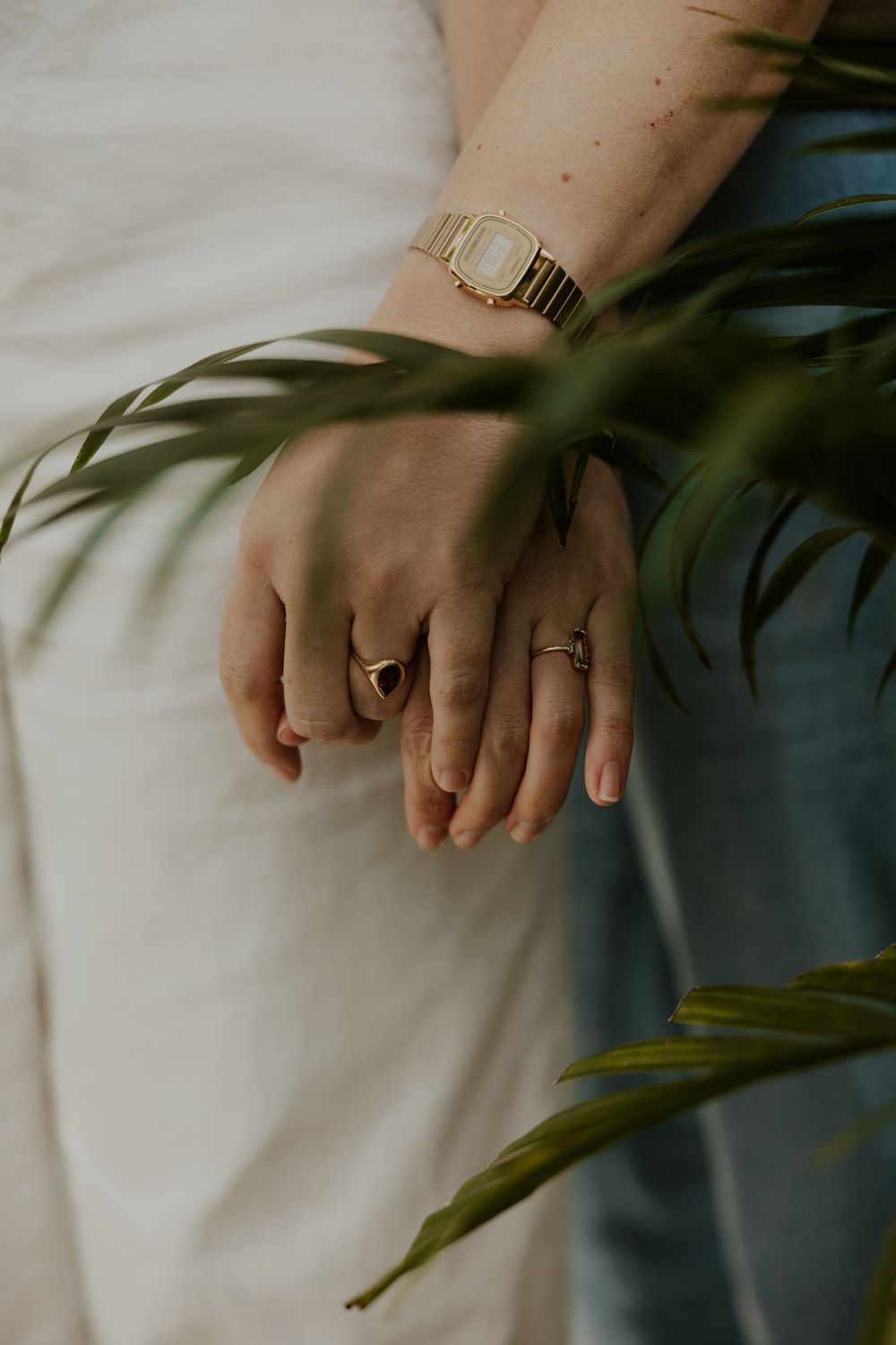Close-up of hands wearing gold rings and watch with green palm leaves in foreground against white fabric background.