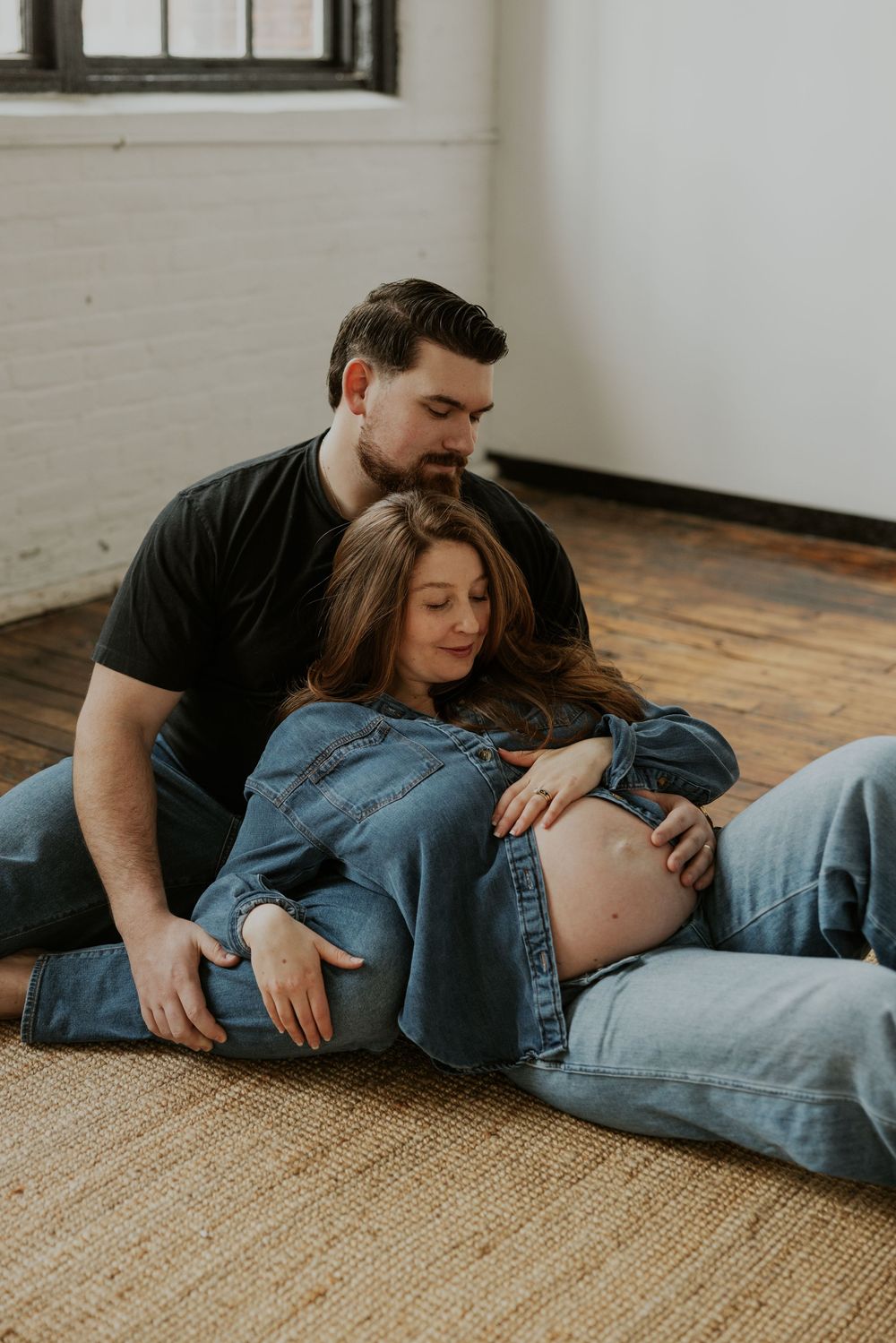 Expectant couple shares intimate moment on hardwood floor with denim and natural light from window.