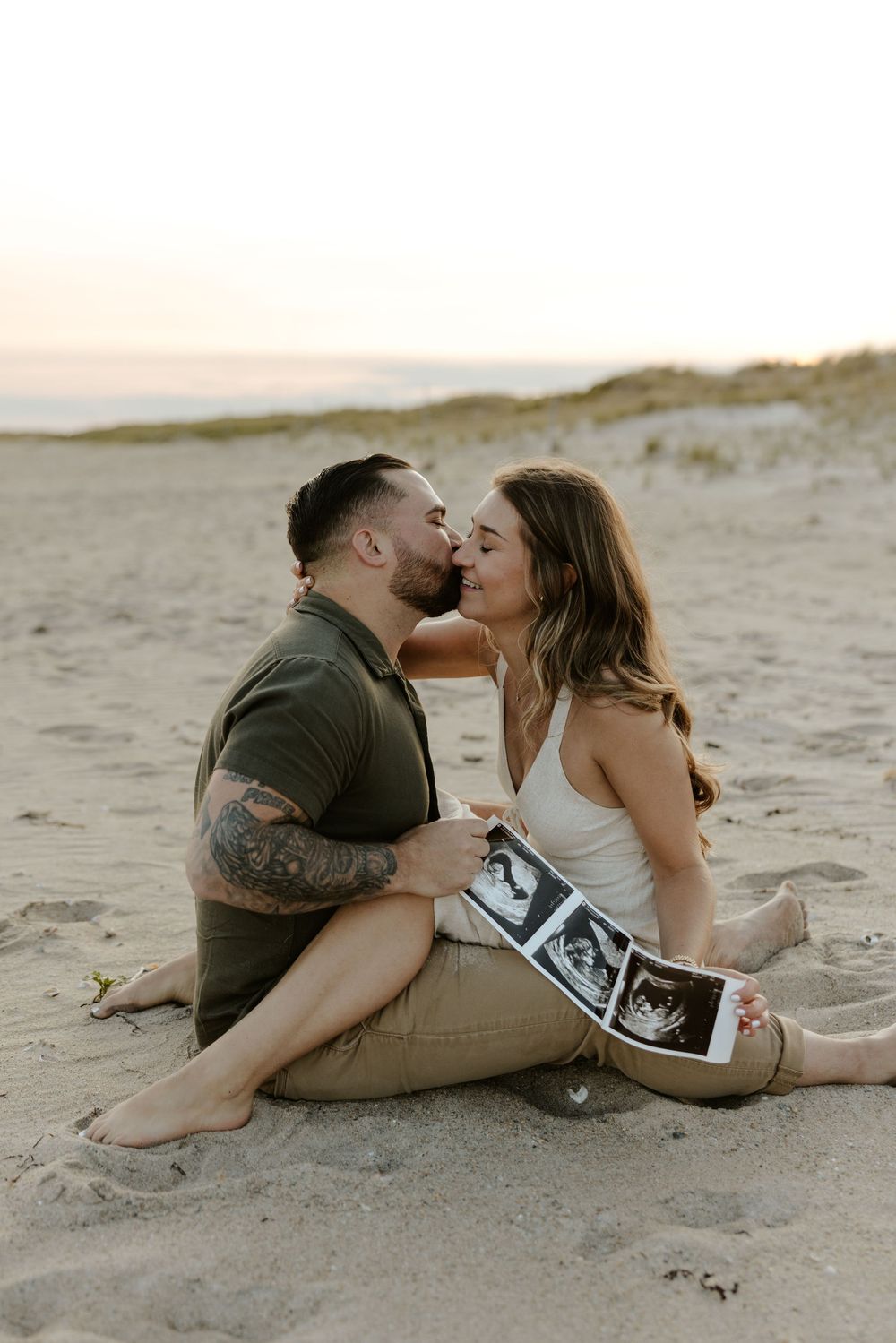 Couple sharing intimate moments on sandy beach while displaying ultrasound photos during sunset pregnancy announcement photoshoot.