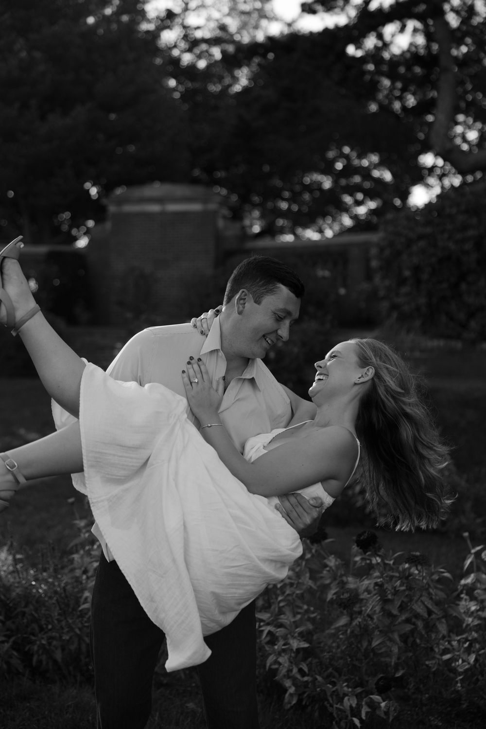A couple in white clothing dips and dances romantically in a garden setting at dusk.