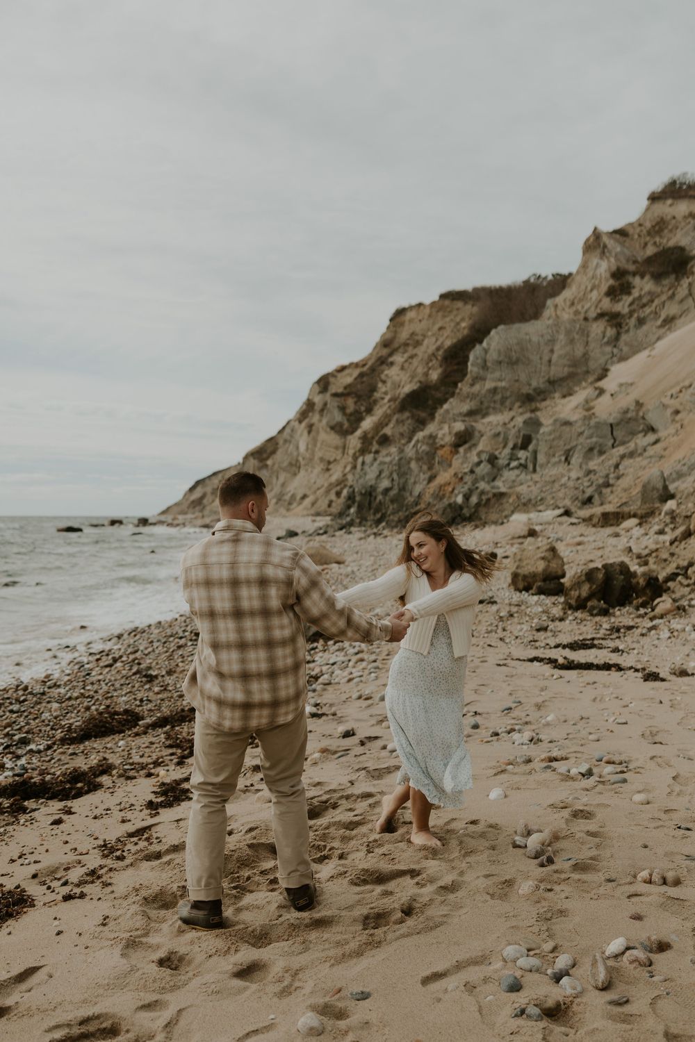 A couple playfully runs and dances together on a rocky beach with dramatic cliffs in the background.
