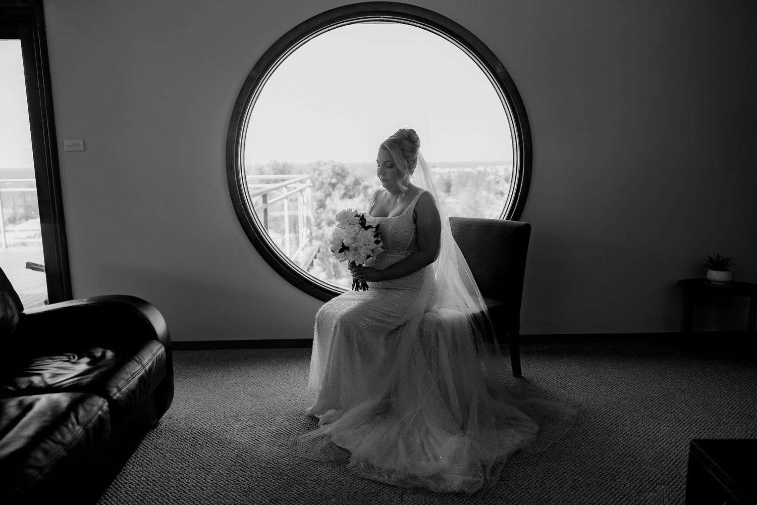 Black and white portrait of a bride sitting by a round window holding a bouquet.