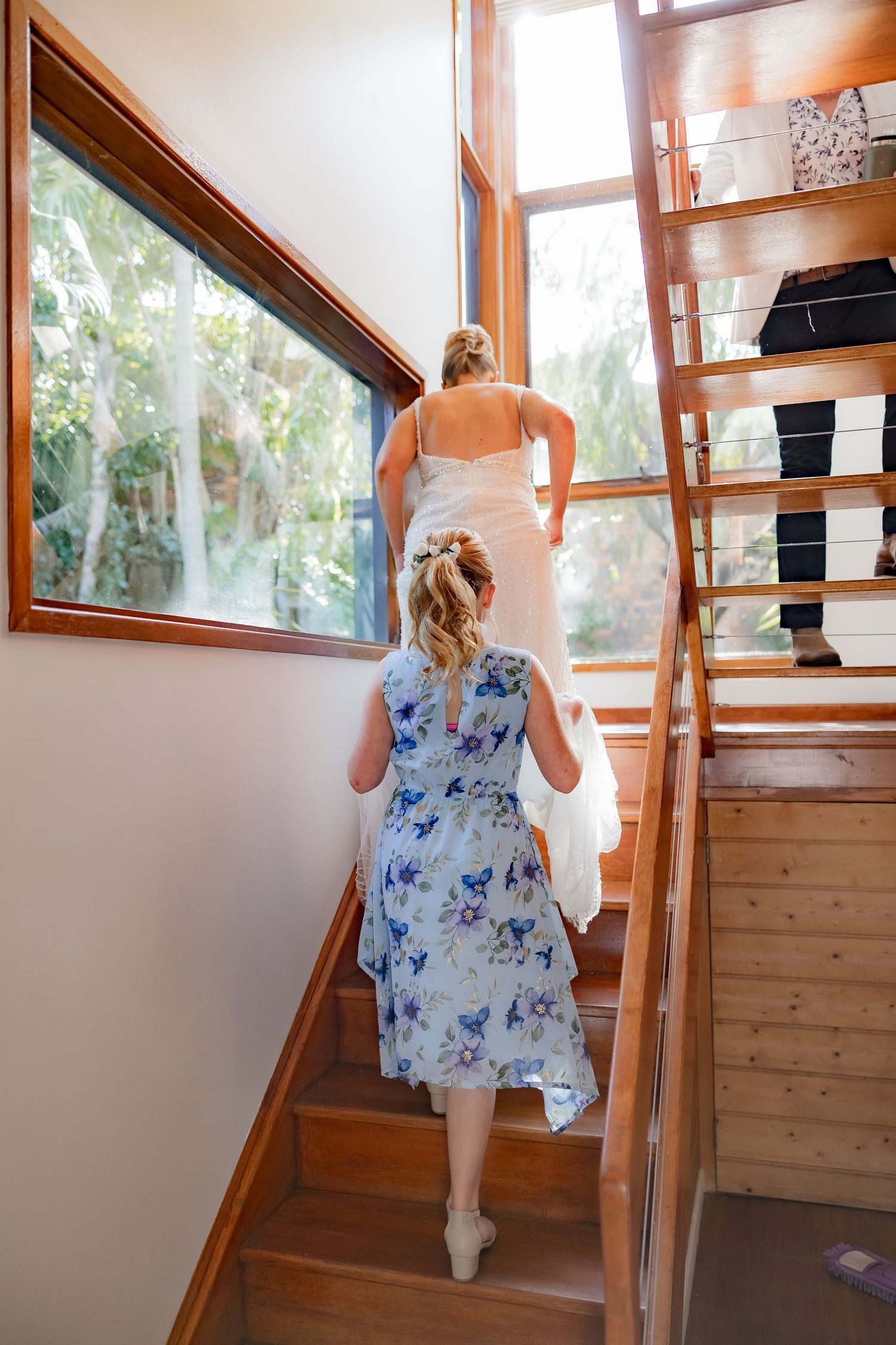 Someone in a floral dress follows another person up wooden stairs in a sunlit room.