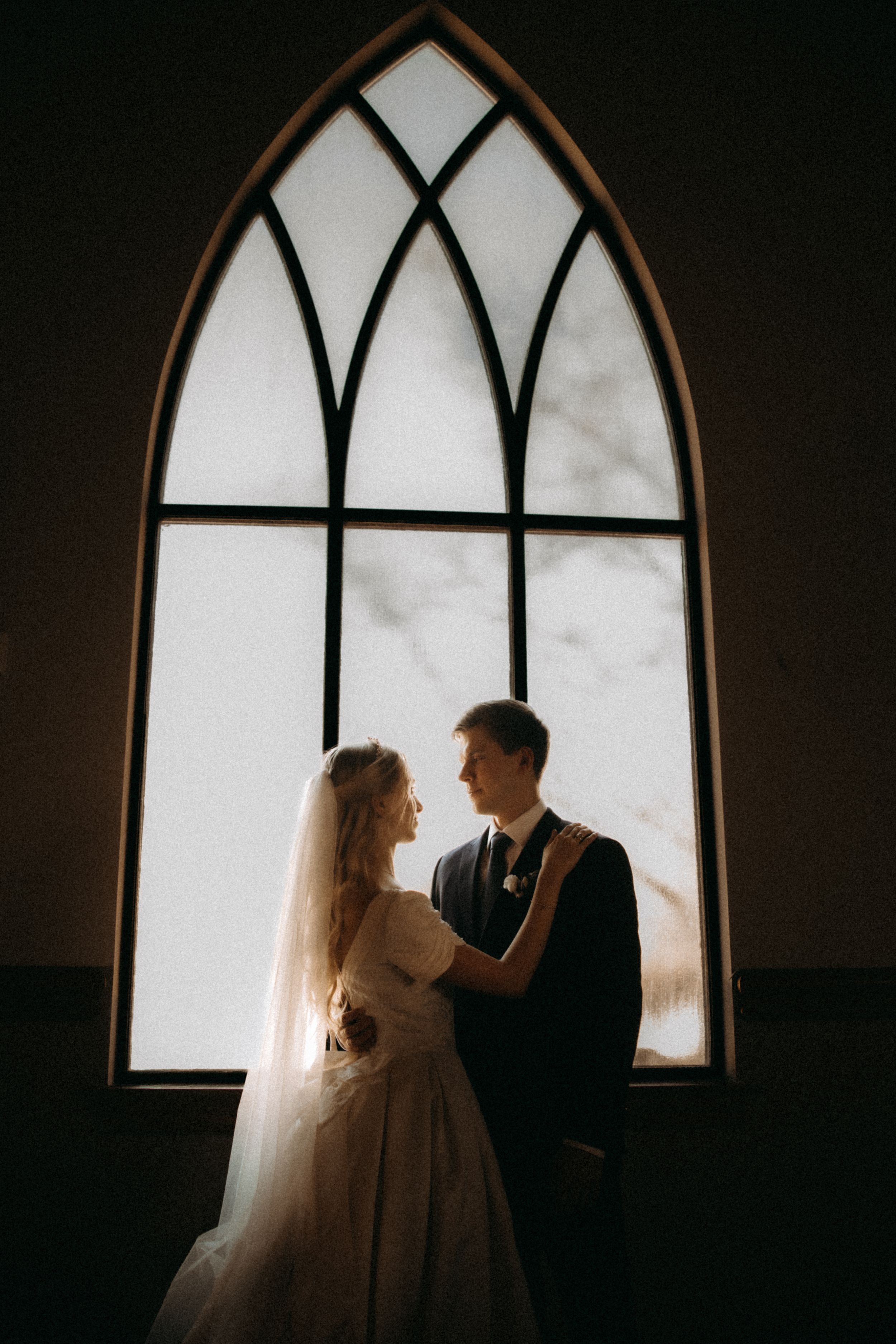 A silhouetted couple in wedding attire stands before a gothic arched window creating a romantic atmosphere.