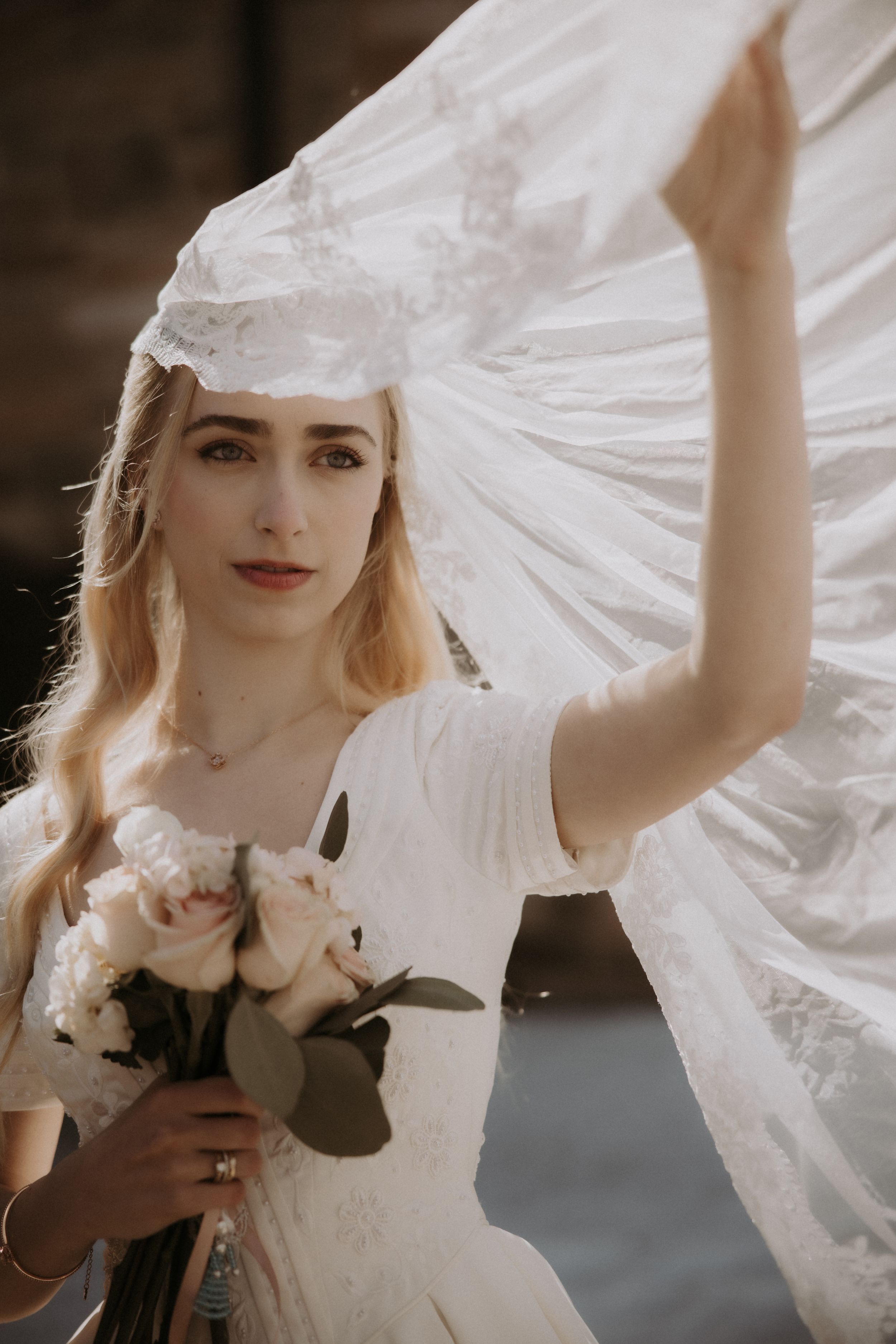 A bride in a white wedding dress holds a bouquet of roses while her delicate veil flows in the breeze.