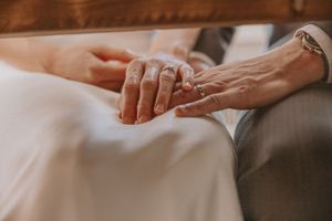 Close-up view of hands wearing wedding rings resting together on white and gray fabric.
