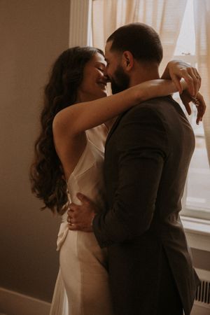 Intimate wedding couple embraces in romantic moody lighting by window.