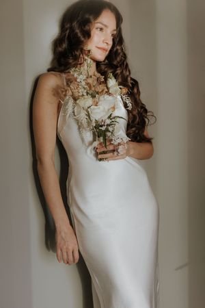Bride in elegant white dress holds delicate bouquet in natural window light.