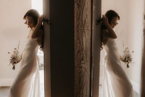 Artistic sepia-toned reflection of bride in wedding dress holding dried flowers by a window.
