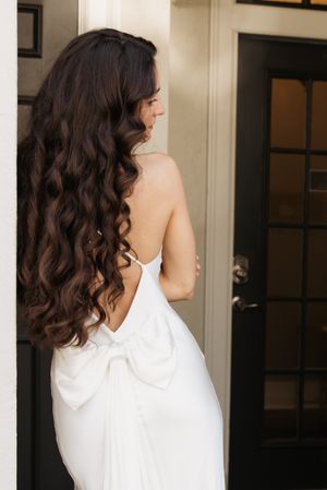 Woman with long dark curly hair wearing a white sleeveless top shown from behind in an indoor setting.