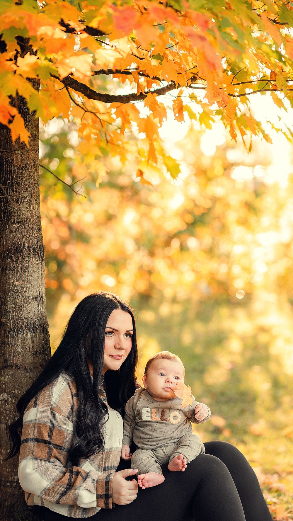 A fall scene with vibrant orange maple leaves as someone sits beneath a tree with an infant in autumn.
