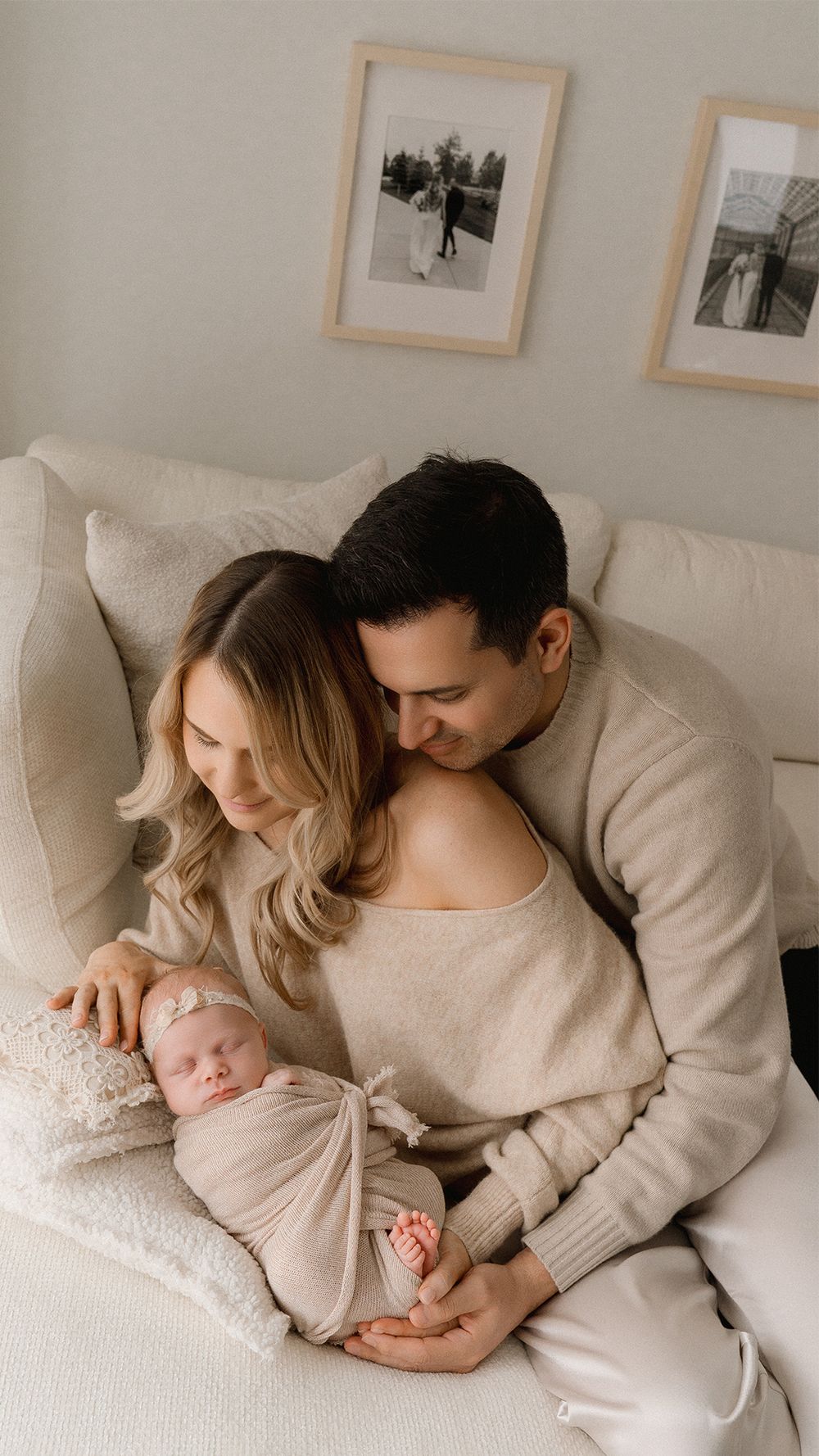 A couple in neutral-toned sweaters cuddles on a white couch with their sleeping newborn baby.