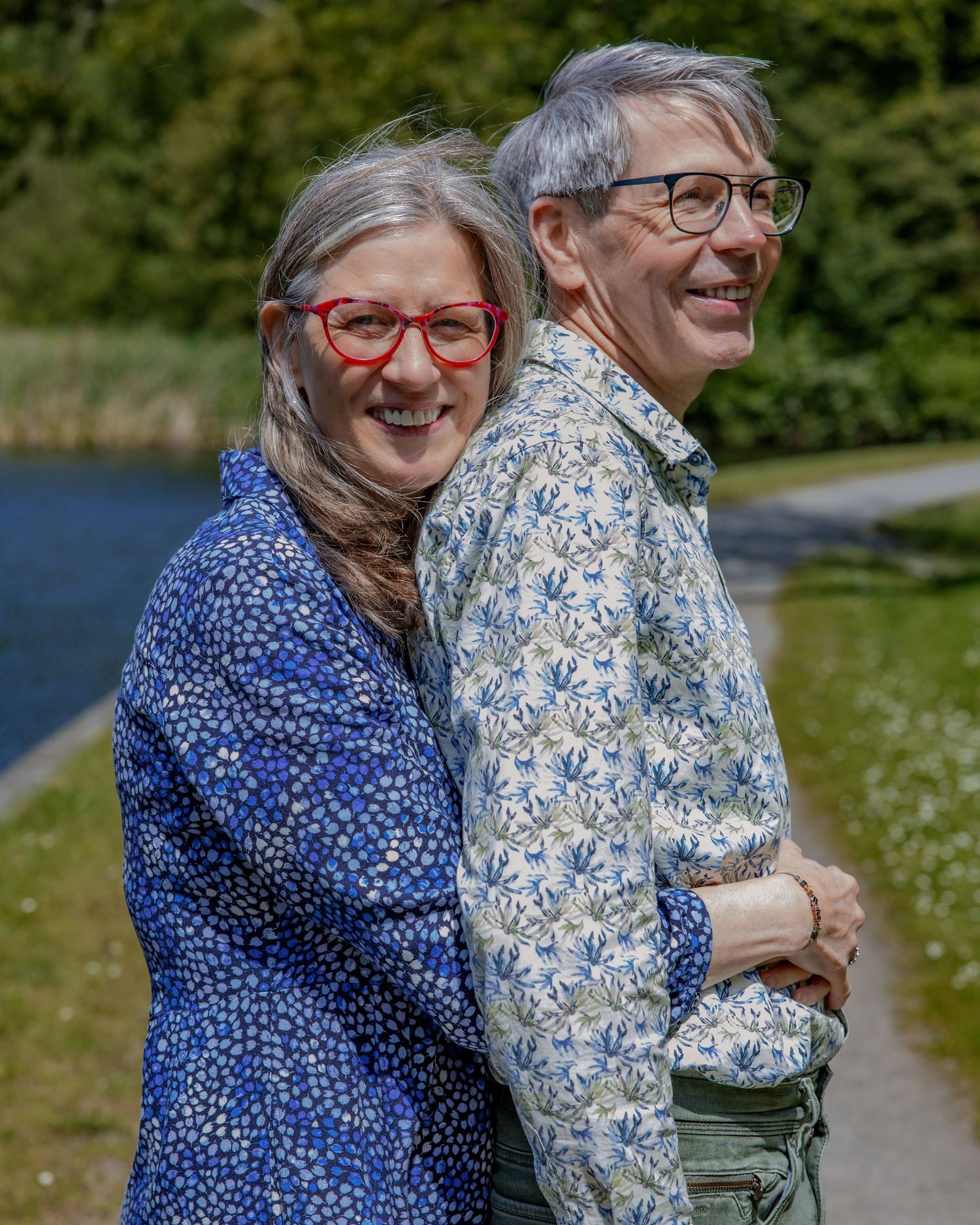 A joyful couple in coordinating floral print shirts embrace while standing outdoors near a lake.

- BU Photography -