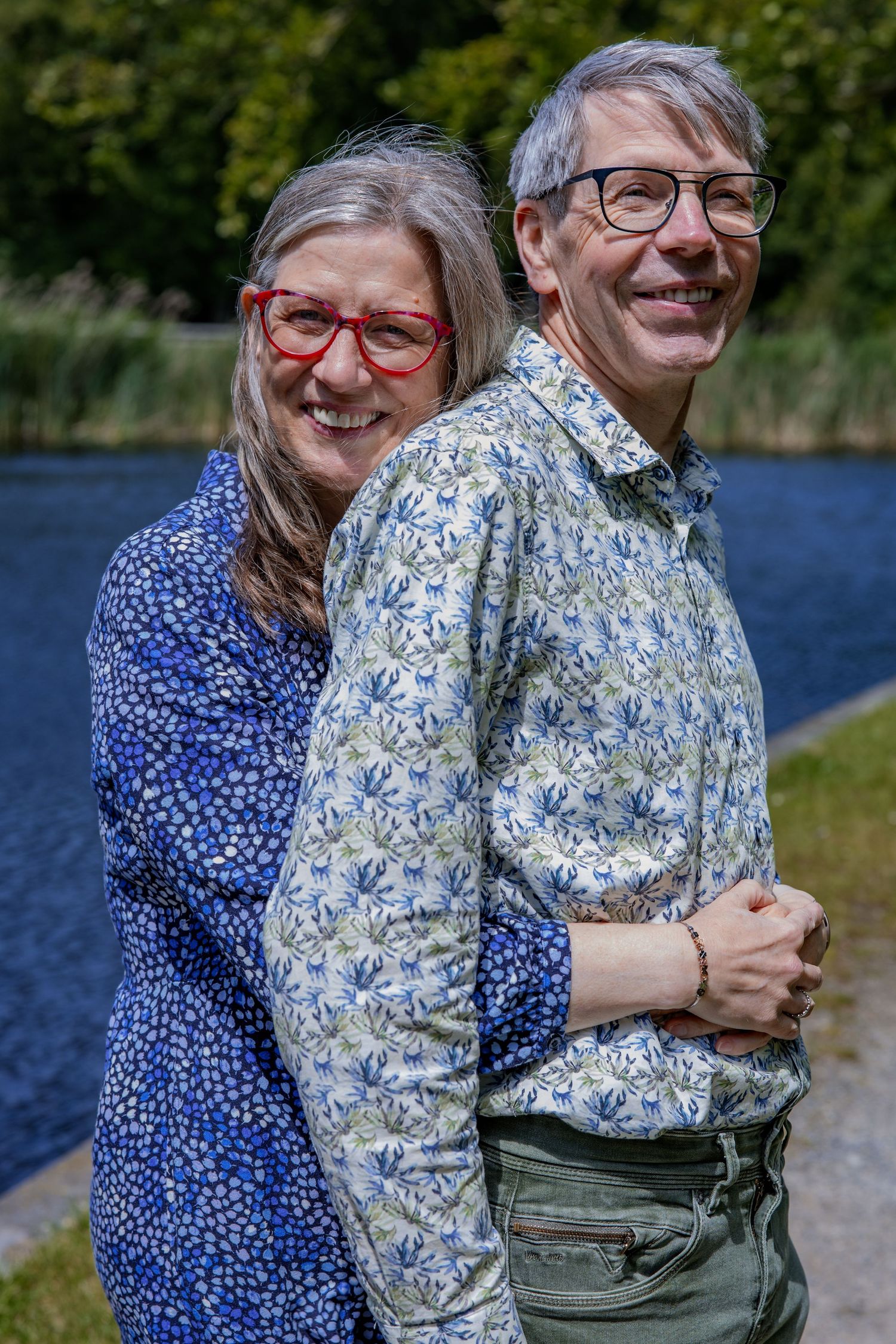 A couple wearing floral patterned shirts embrace and smile while standing by a lake on a sunny day.

- BU Photography -