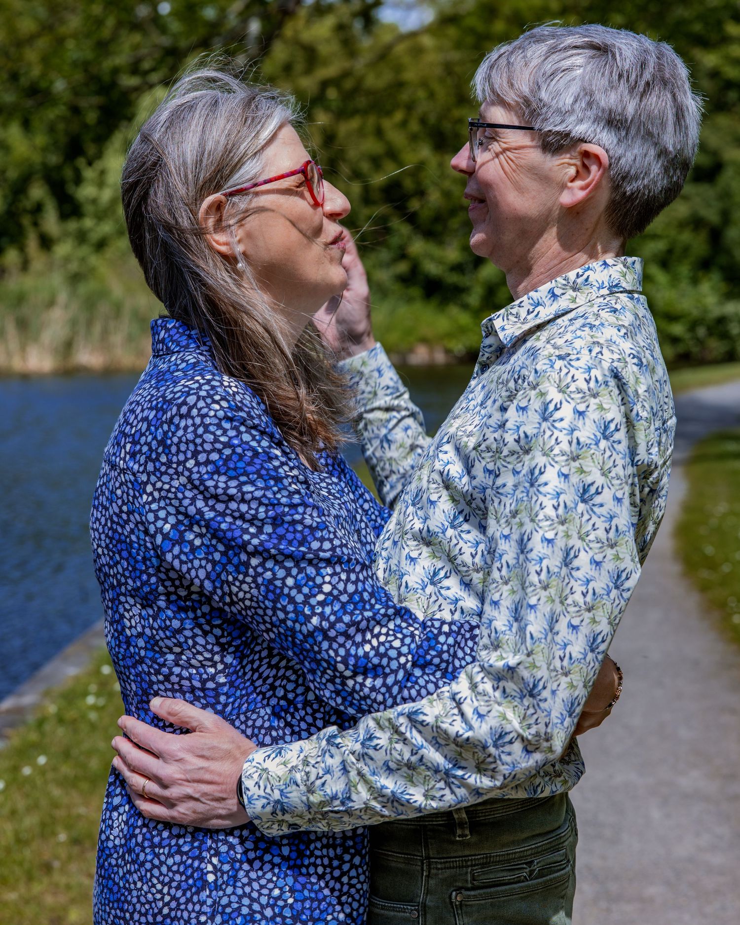 Two people in floral print shirts share an intimate moment while embracing on a path near a lake on a sunny day.

- BU Photography -