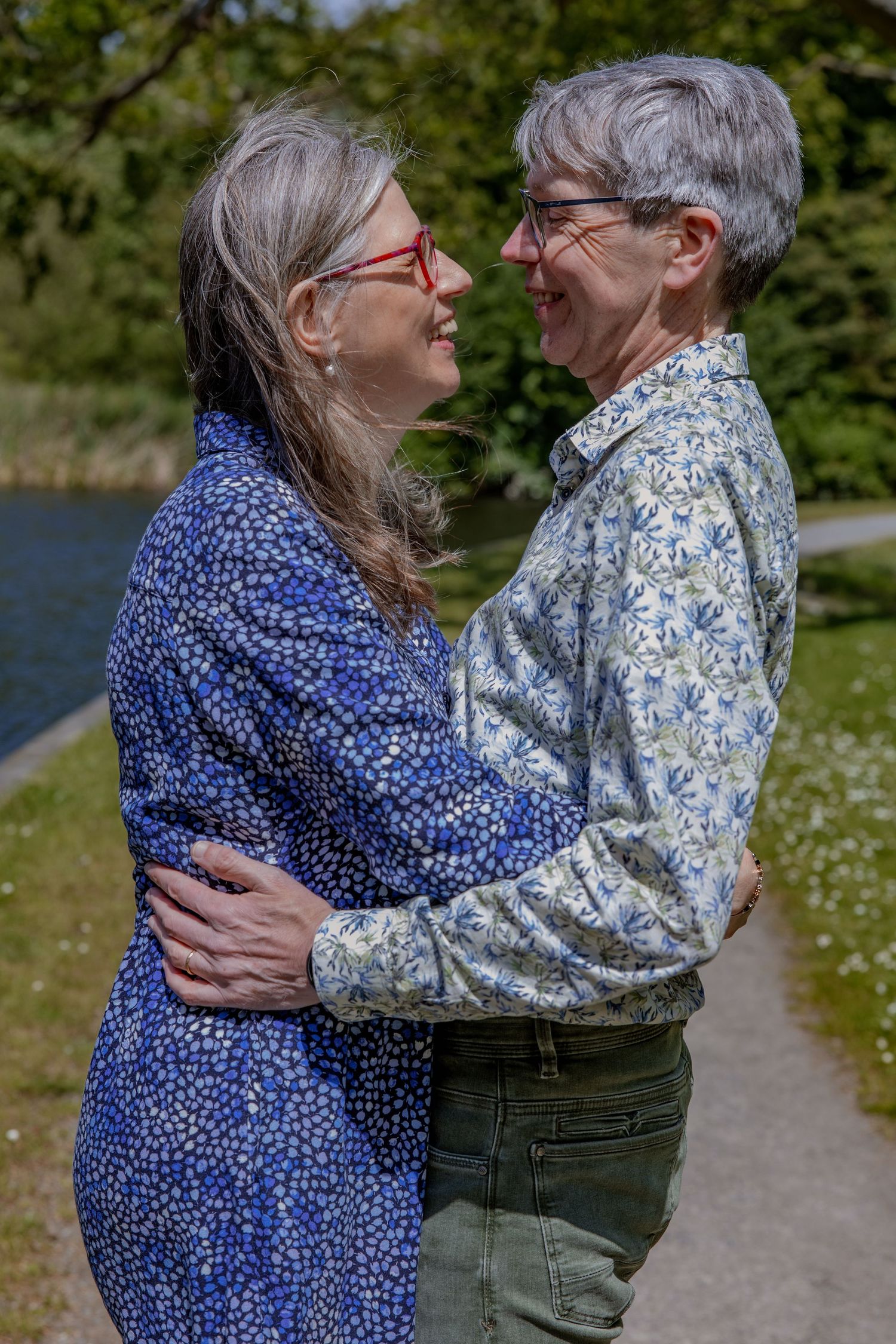 A couple in blue and floral print shirts share an intimate moment on a path by a lake.

- BU Photography -