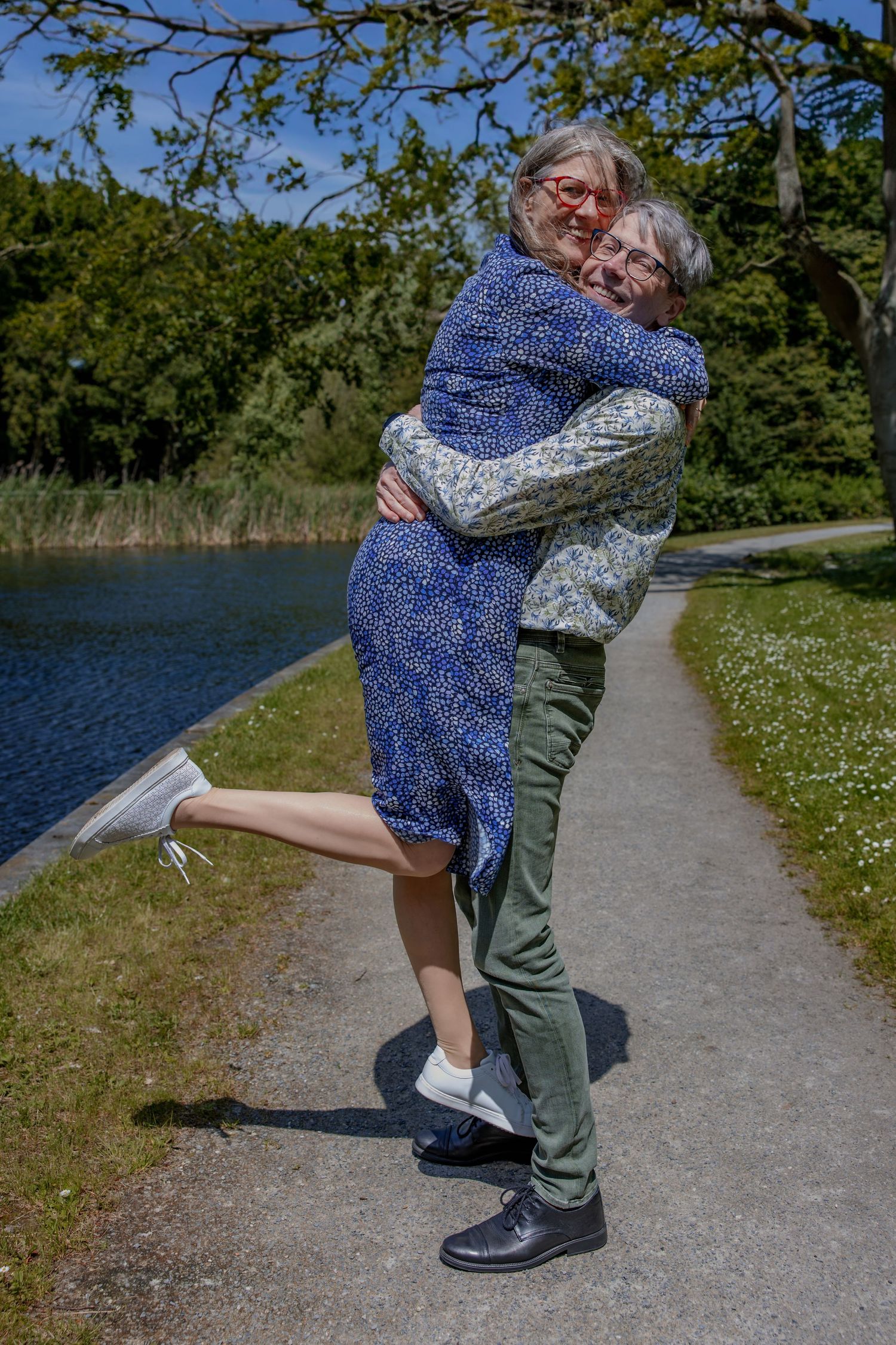 Two people share a joyful embrace on a sunny path by a lake, with green trees and grass in the background.

- BU Photography -