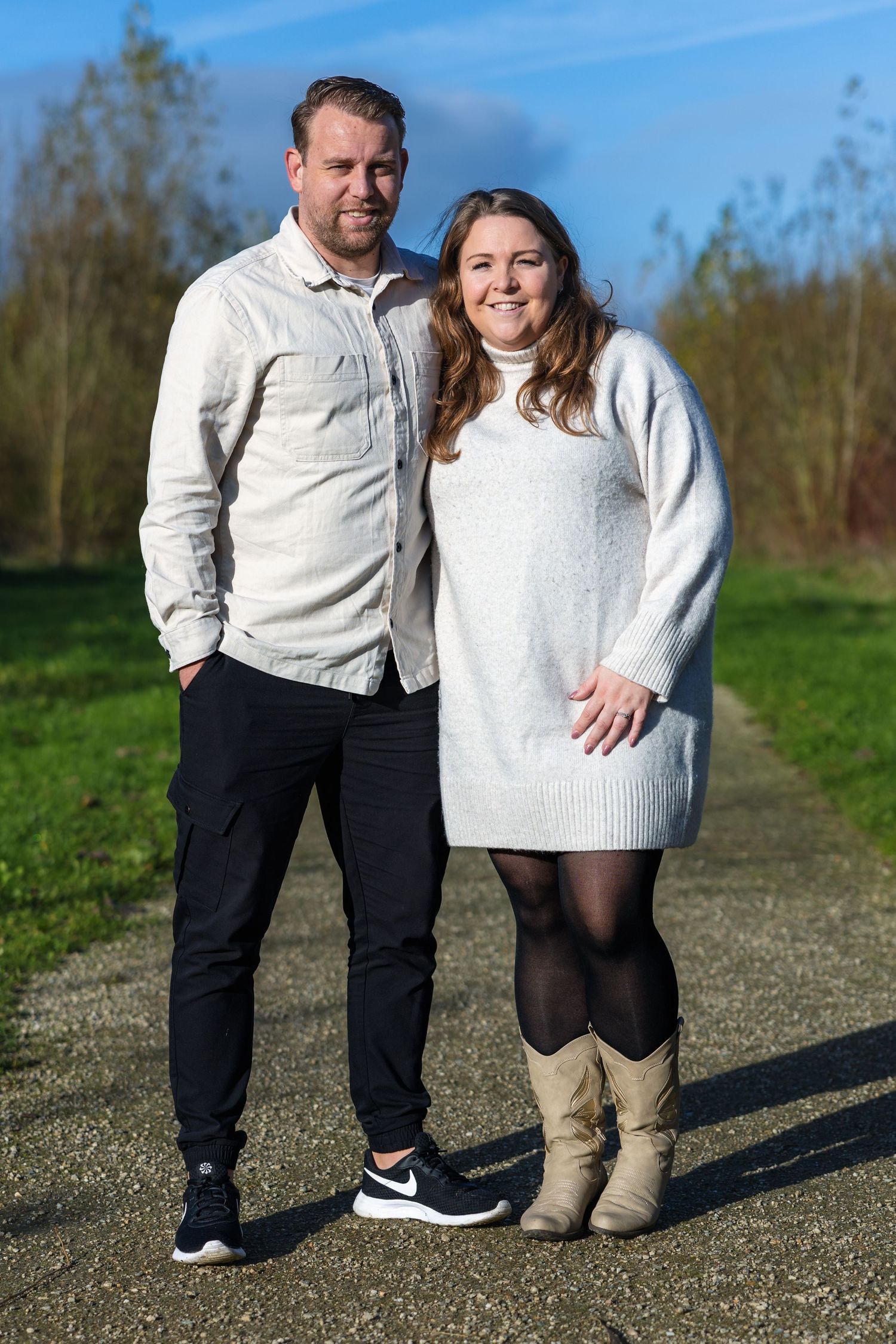 A couple wearing white sweaters pose together on a dirt path on a sunny day with trees in the background.