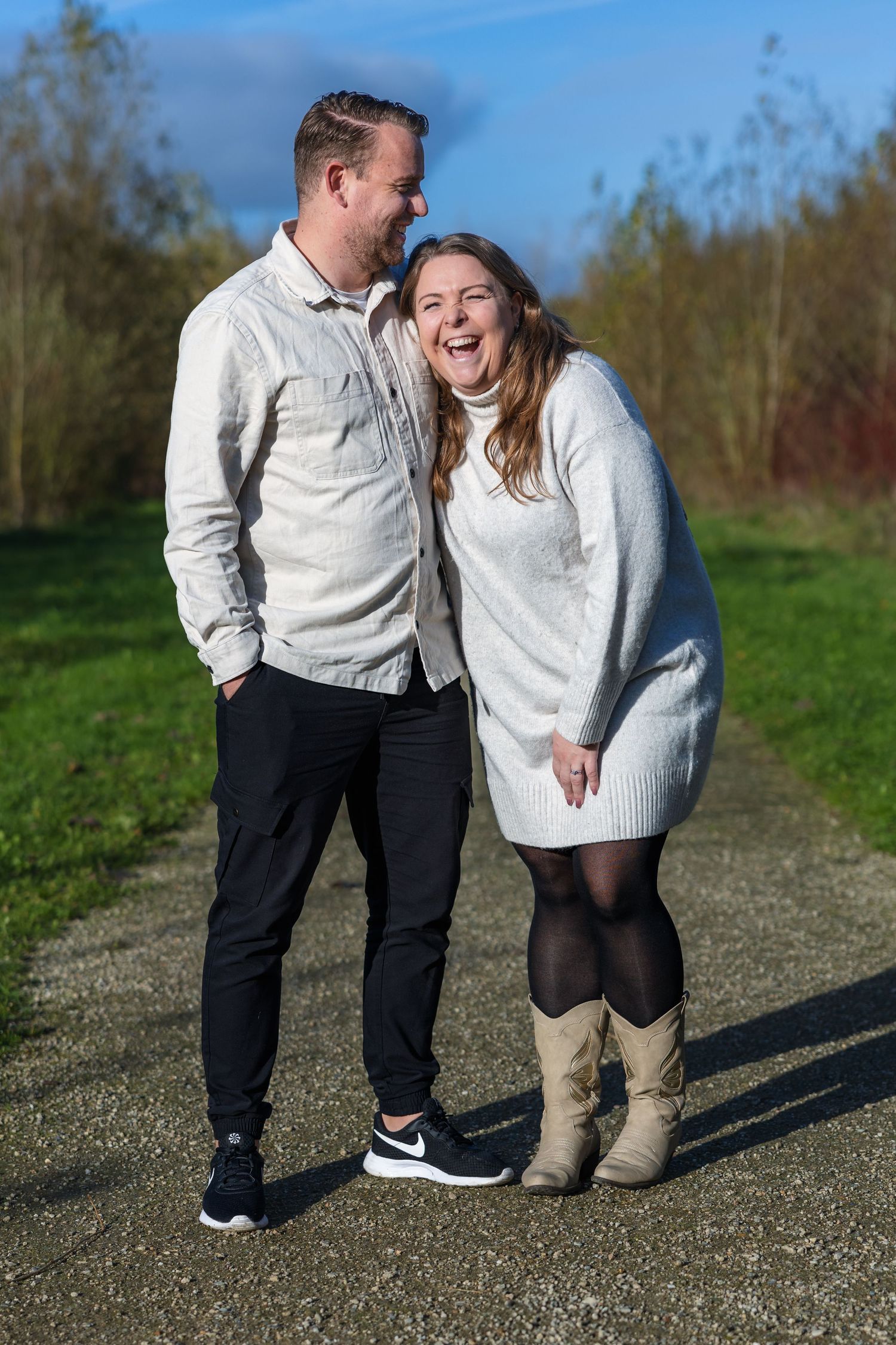A couple in coordinating light sweaters laugh together on a sunny path with trees in the background.