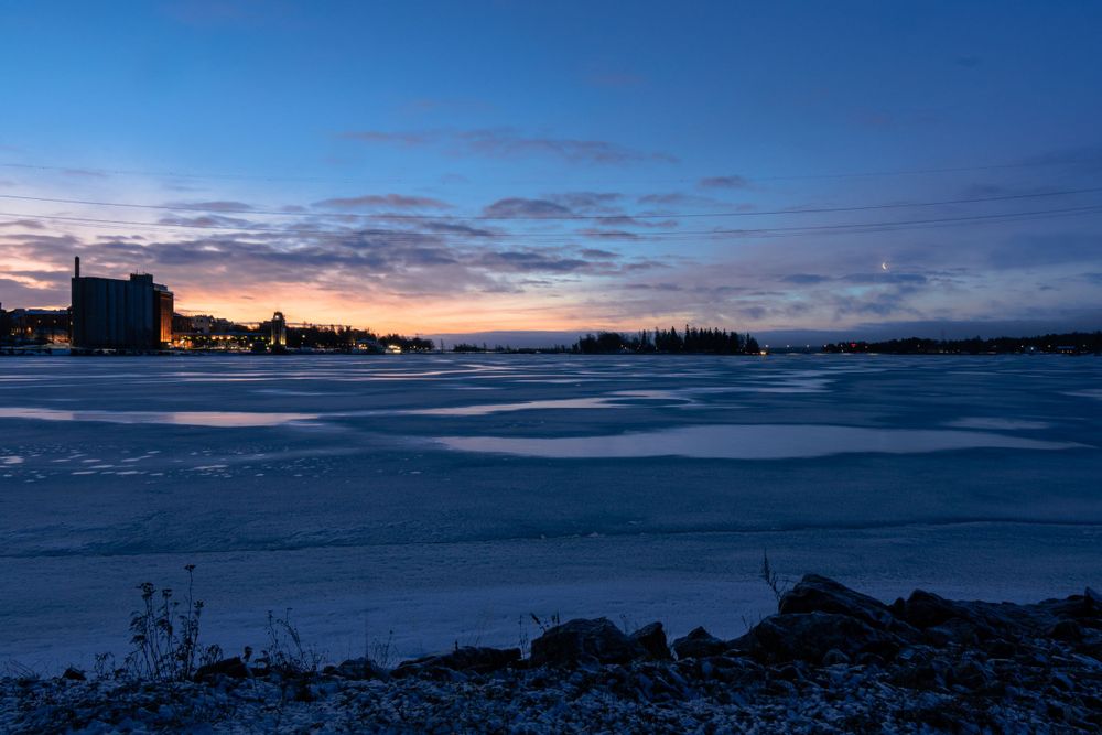 Twilight sky reflects over a frozen lake landscape with silhouetted buildings along the snowy shore at dusk.