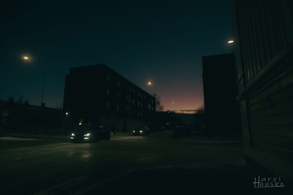 Dark urban street at dusk with silhouettes of buildings against a moody twilight sky and distant street lights glowing.