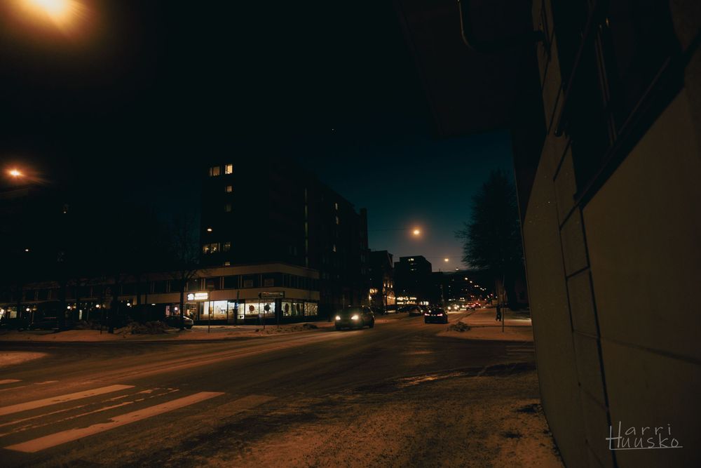 Dark urban street at night with glowing streetlights and buildings silhouetted against a dim sky.