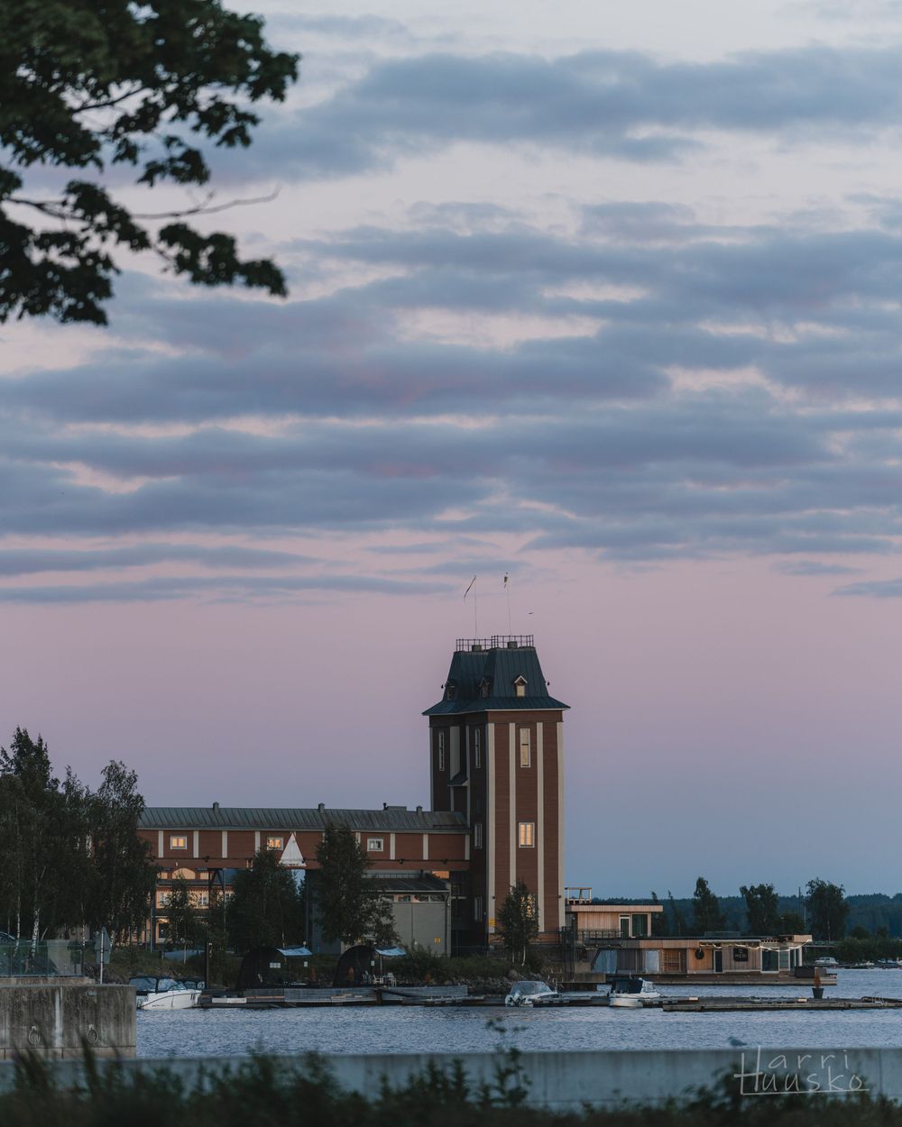 Historic tower building silhouetted against pink and purple sunset clouds near a waterfront.