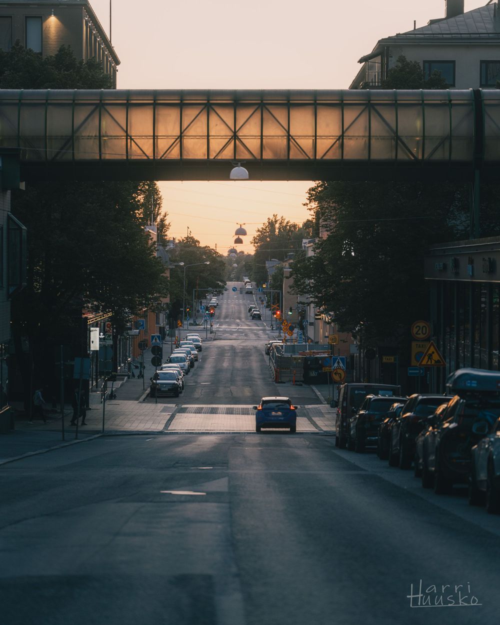 Urban street scene with a pedestrian bridge crossing between buildings at sunset.