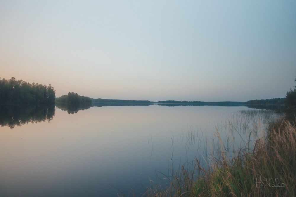 Tranquil sunset over a calm lake surrounded by forested shoreline.