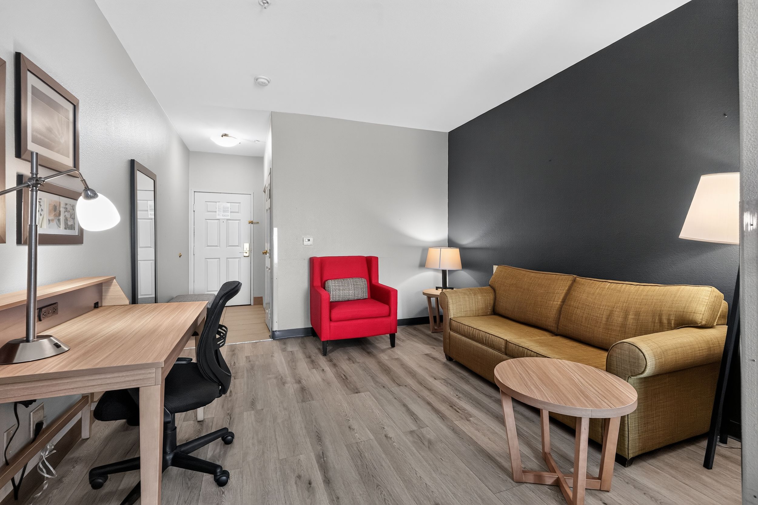 Modern hotel suite with gold sofa, red accent chair, wood desk, and dark feature wall with warm lighting.