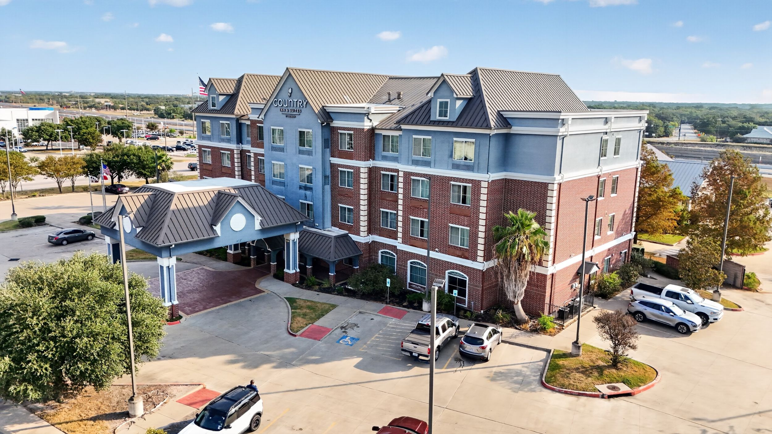 Aerial view of a brick hotel with blue accents, porte-cochere entrance, and parking lot surrounded by trees.