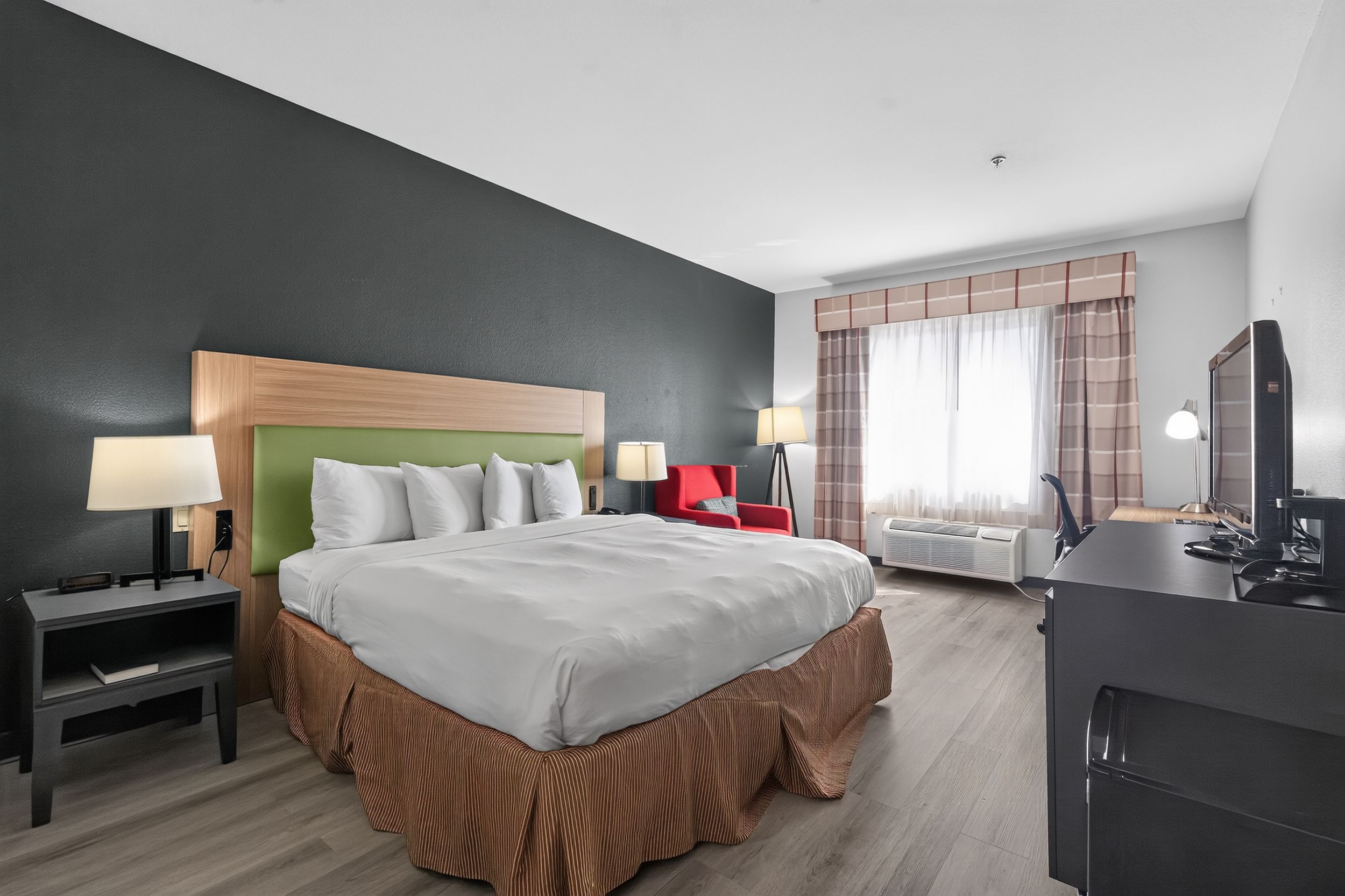 Modern hotel room with king bed, green headboard, red plaid curtains, hardwood floors, and TV on dark wood dresser.