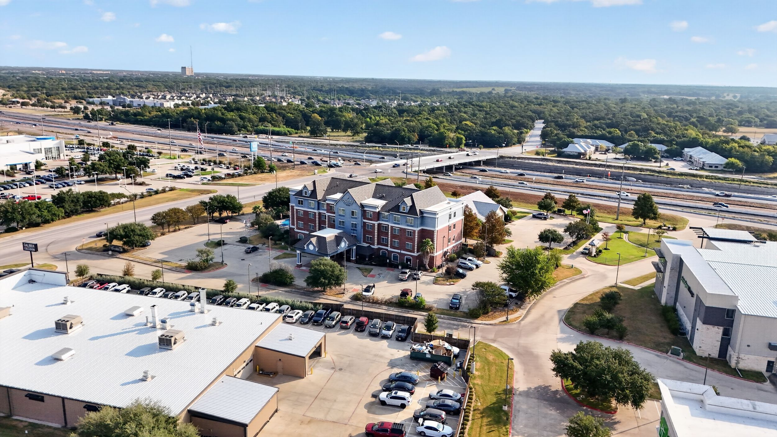Aerial view of a historic brick building with parking lot, highway, and lush green trees in the background.