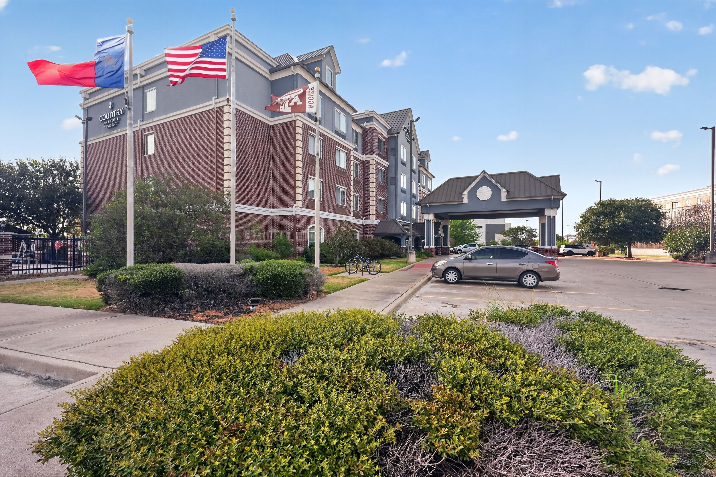 Exterior view of a brick hotel with American and Texas flags, landscaped shrubs, and a covered entrance canopy.