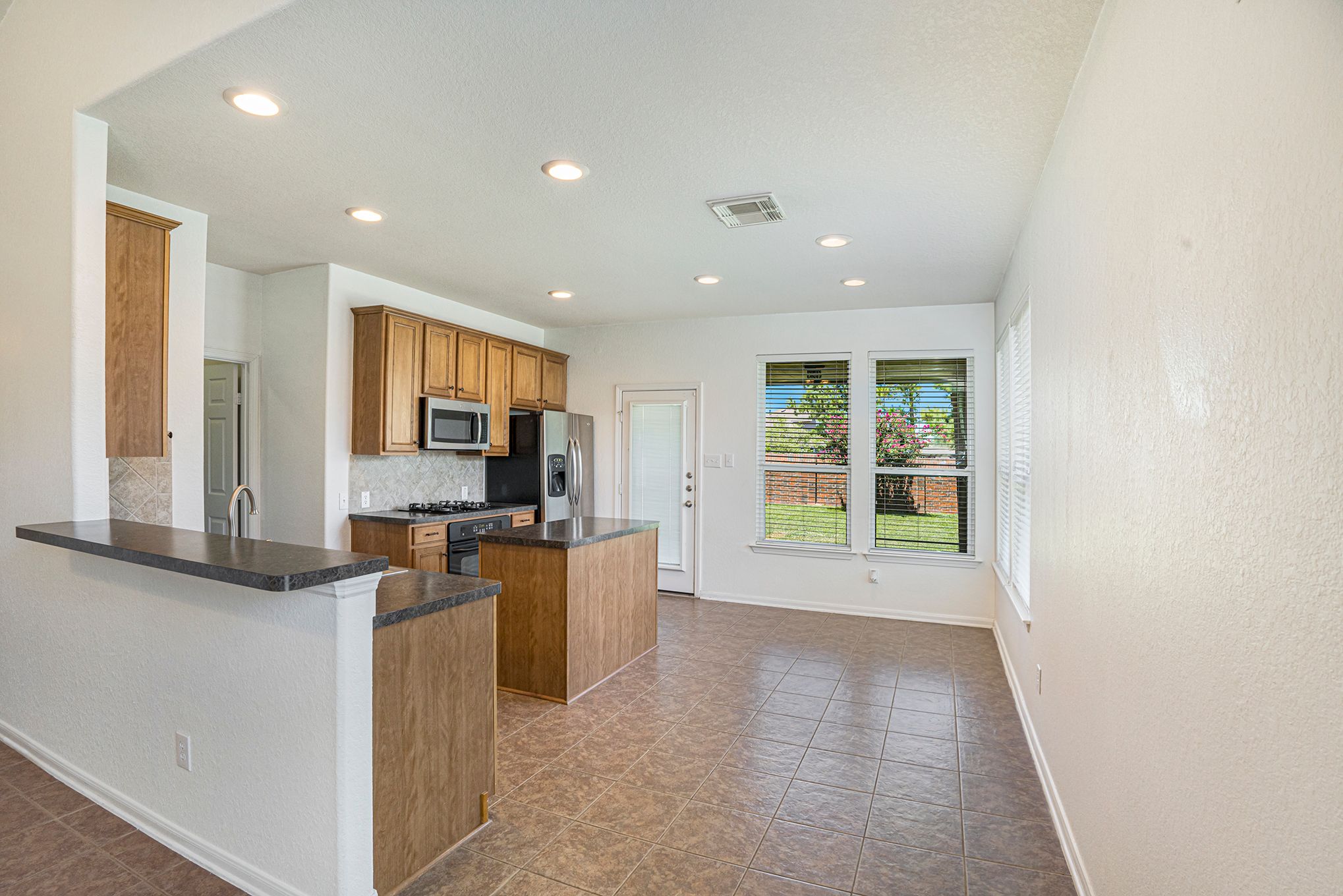 Modern kitchen with wood cabinets, dark countertops, and stainless appliances opens to bright living area with backyard view.