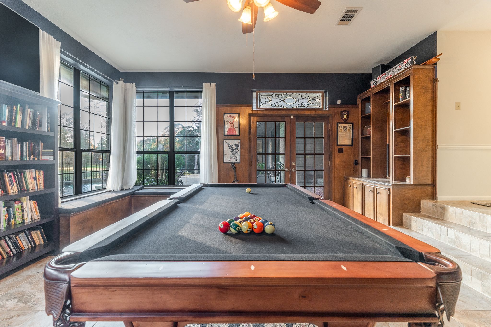 A panoramic view of a game room interior with pool tables, bookshelves, and ceiling fans in warm lighting.