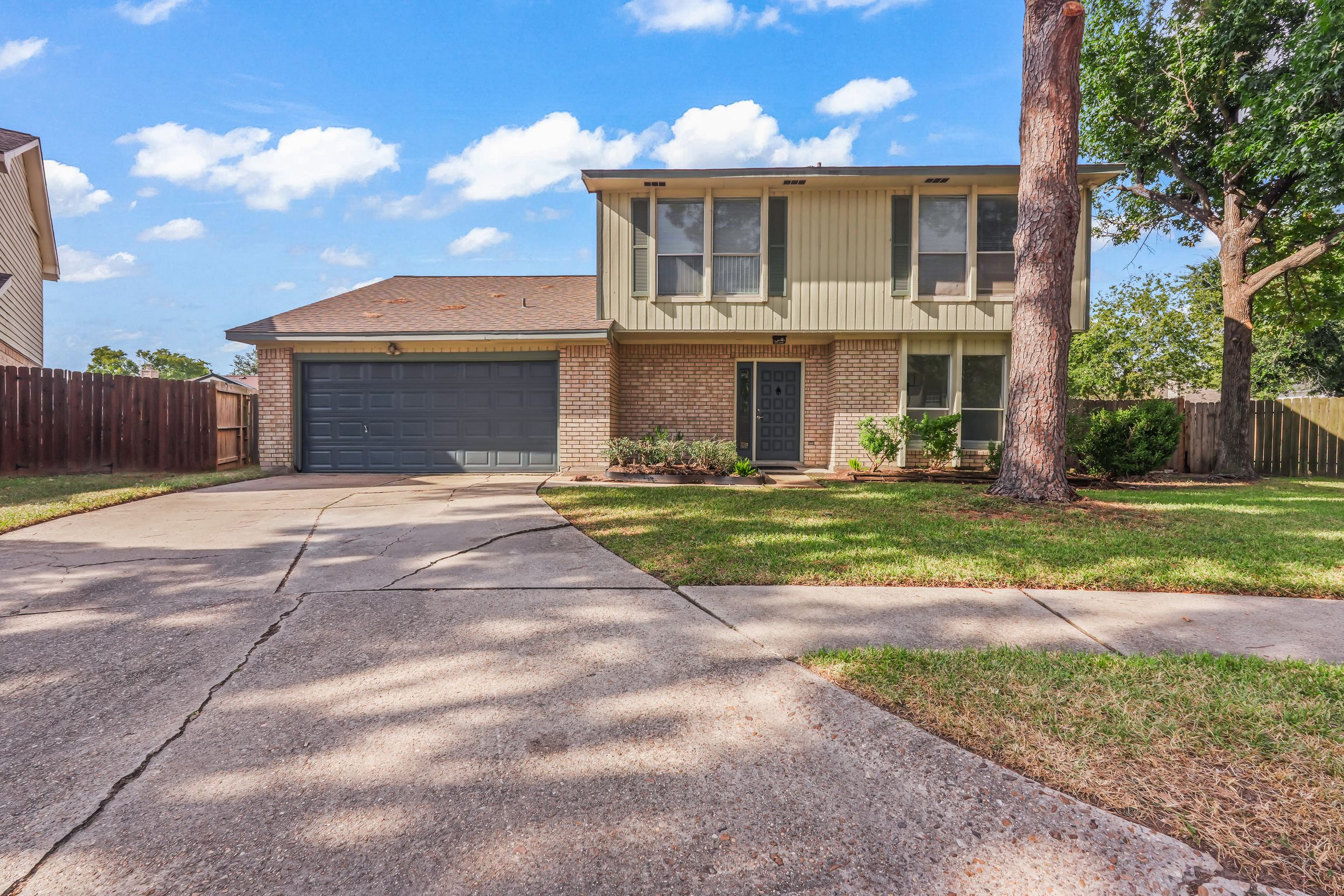 Two-story brick home with green lawn, attached two-car garage, and mature trees on a sunny day.